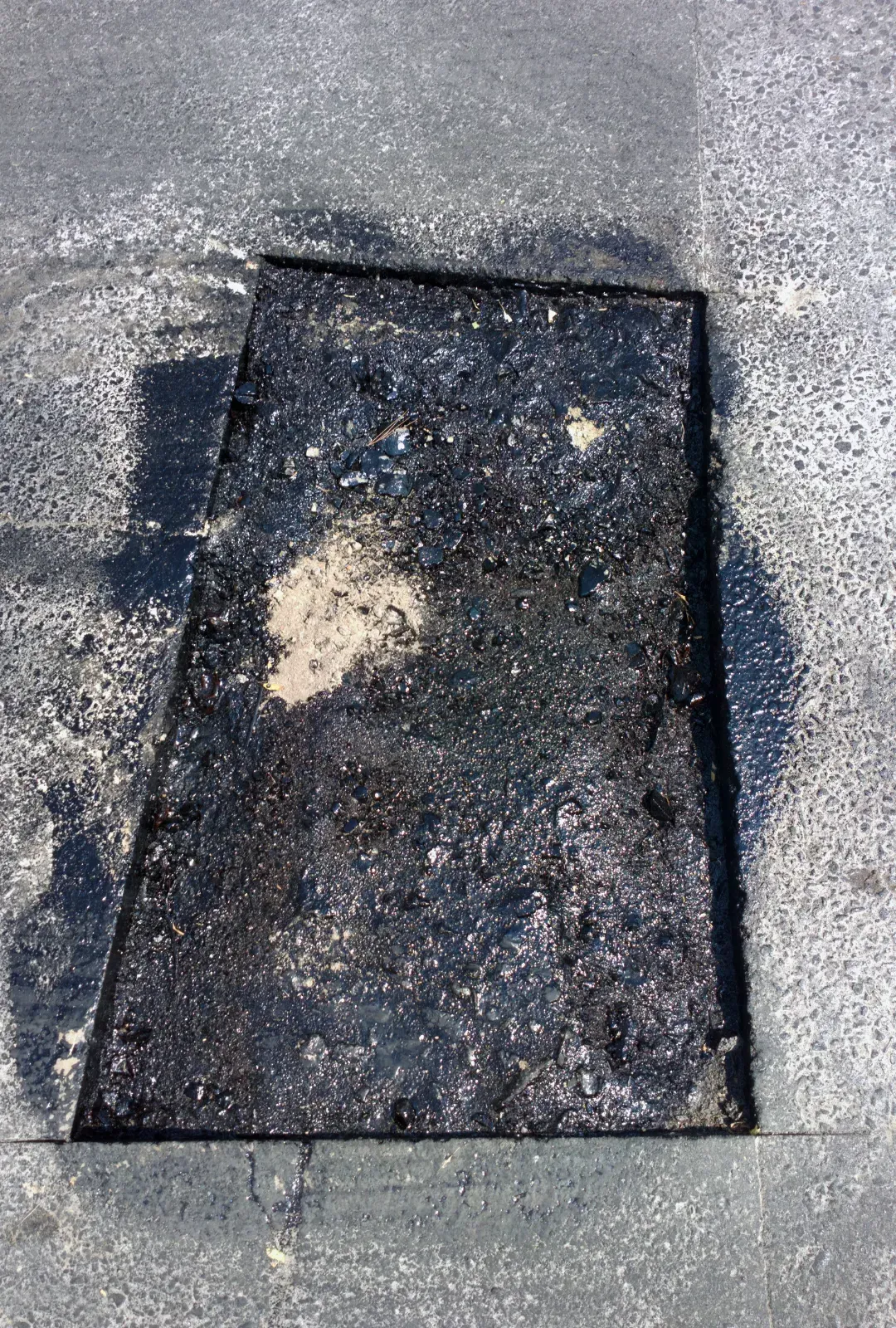 A rectangular patch of fresh black asphalt filling a repair cut in an existing gray road surface.