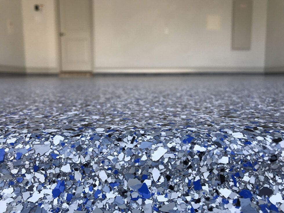 A close-up view of a garage floor with a blue, gray, and white flake epoxy finish, showing a sharp, clean edge.