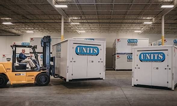 A warehouse worker operates a yellow forklift to move a white storage container marked with the blue 