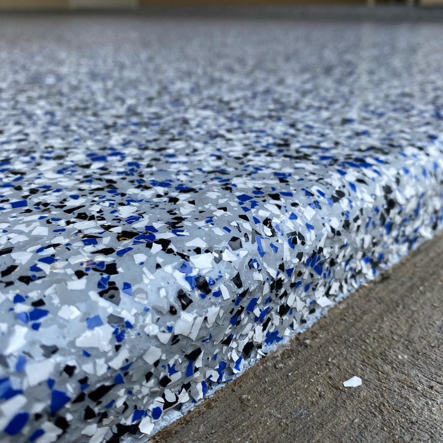 A garage floor coated with blue, white, and black paint flakes, showing the textured edge where it meets plain concrete.