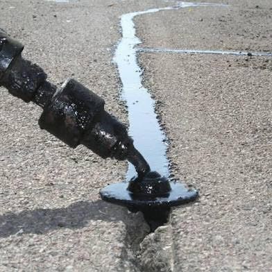 A nozzle applies hot, black liquid sealant to fill a crack in a textured asphalt surface.