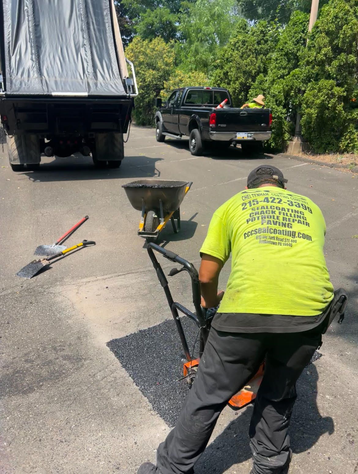 A worker in a neon yellow shirt uses a tool to smooth fresh asphalt in a driveway, with a dump truck and pickup nearby.