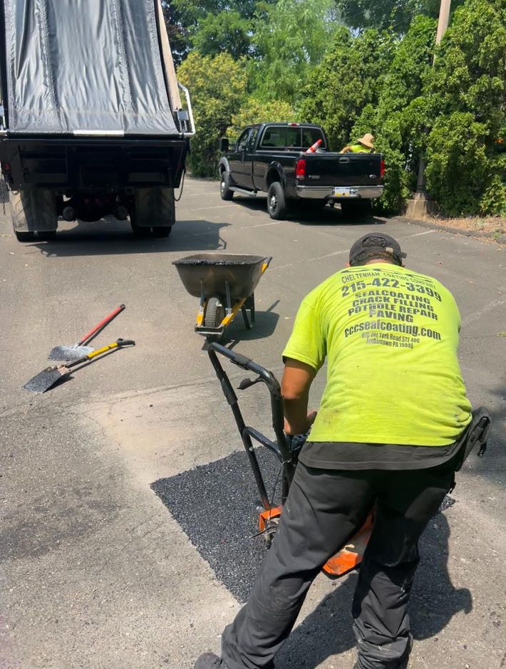 A worker in a neon yellow shirt uses a tool to smooth fresh asphalt in a driveway with a truck and trailer nearby.