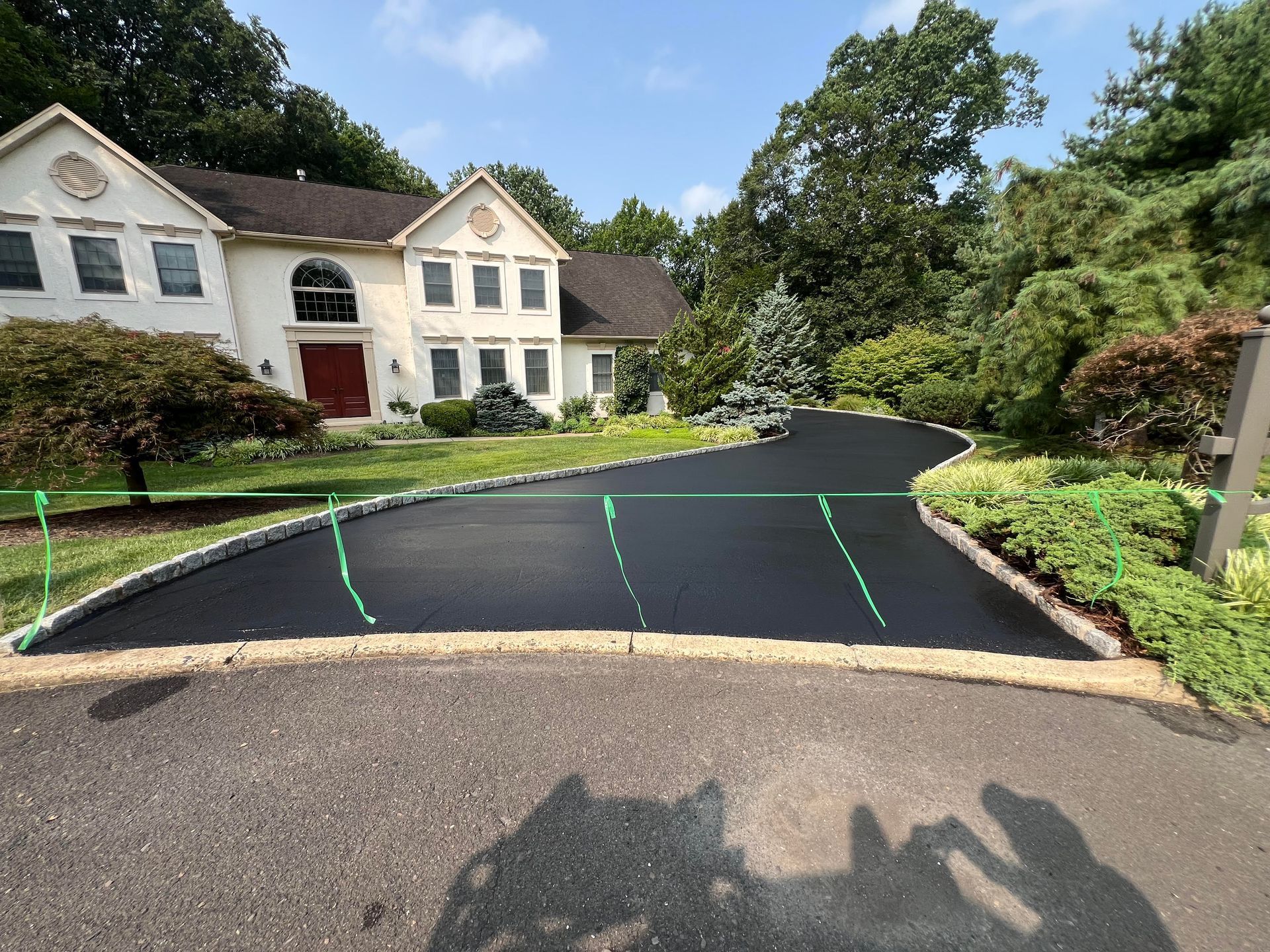 A freshly paved black asphalt driveway leads to a large, light-colored suburban house surrounded by green trees and shrubs.