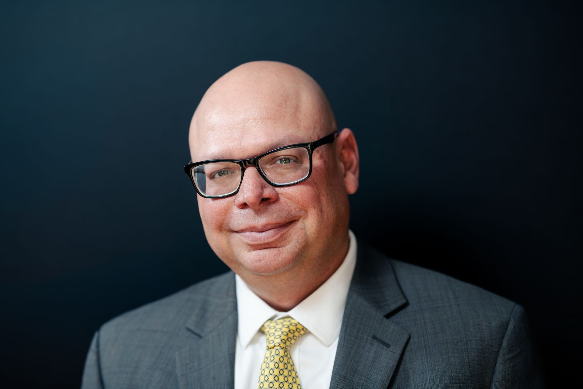 Man in a navy blazer and white shirt smiles at the camera, against a gray backdrop.