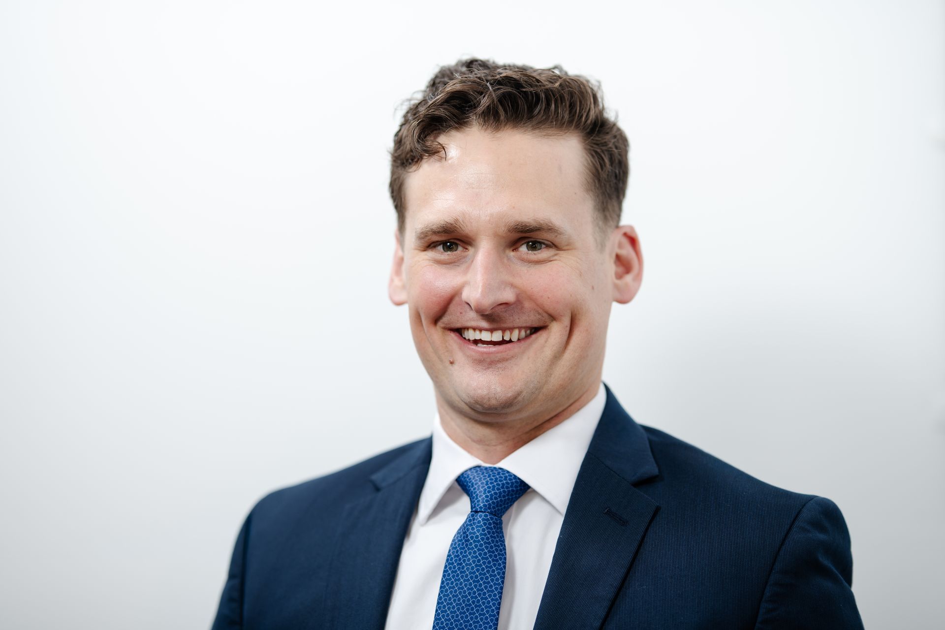 Smiling person in a white shirt and patterned tie against a light wall