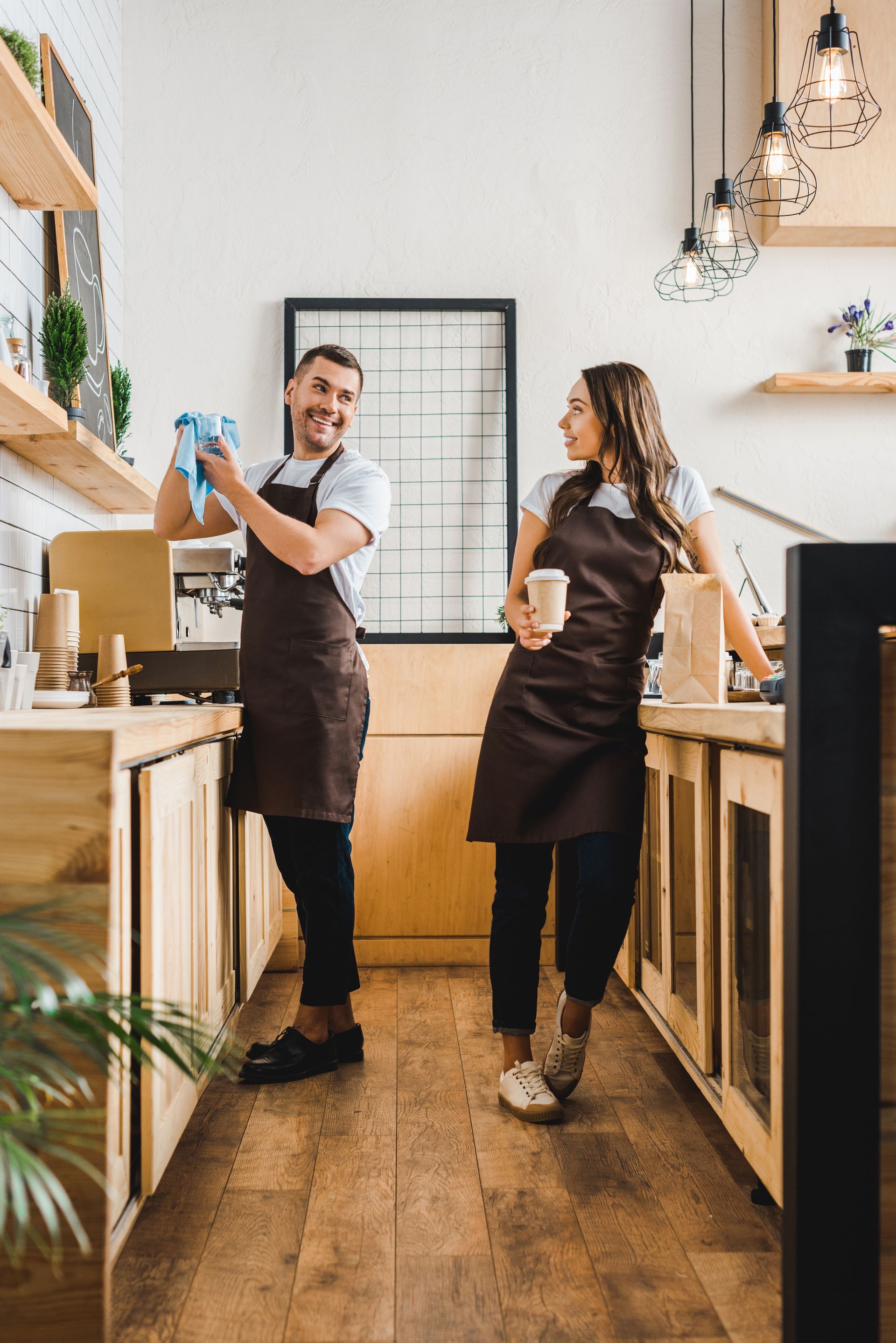 Two baristas in brown aprons smiling in a bright café kitchen, preparing drinks behind the counter.