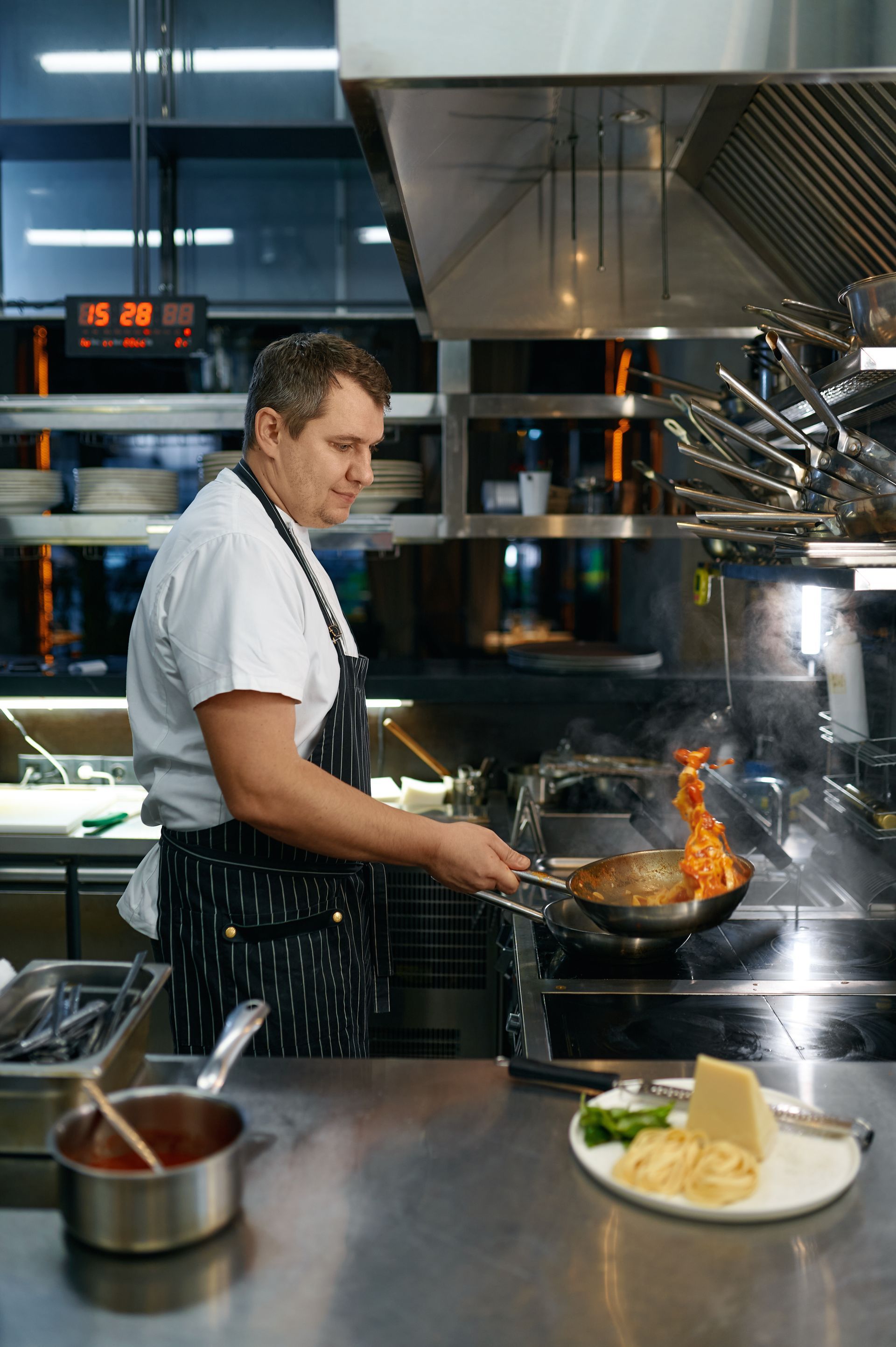 A chef wearing a white uniform and patterned apron flambés food in a pan in a professional kitchen.
