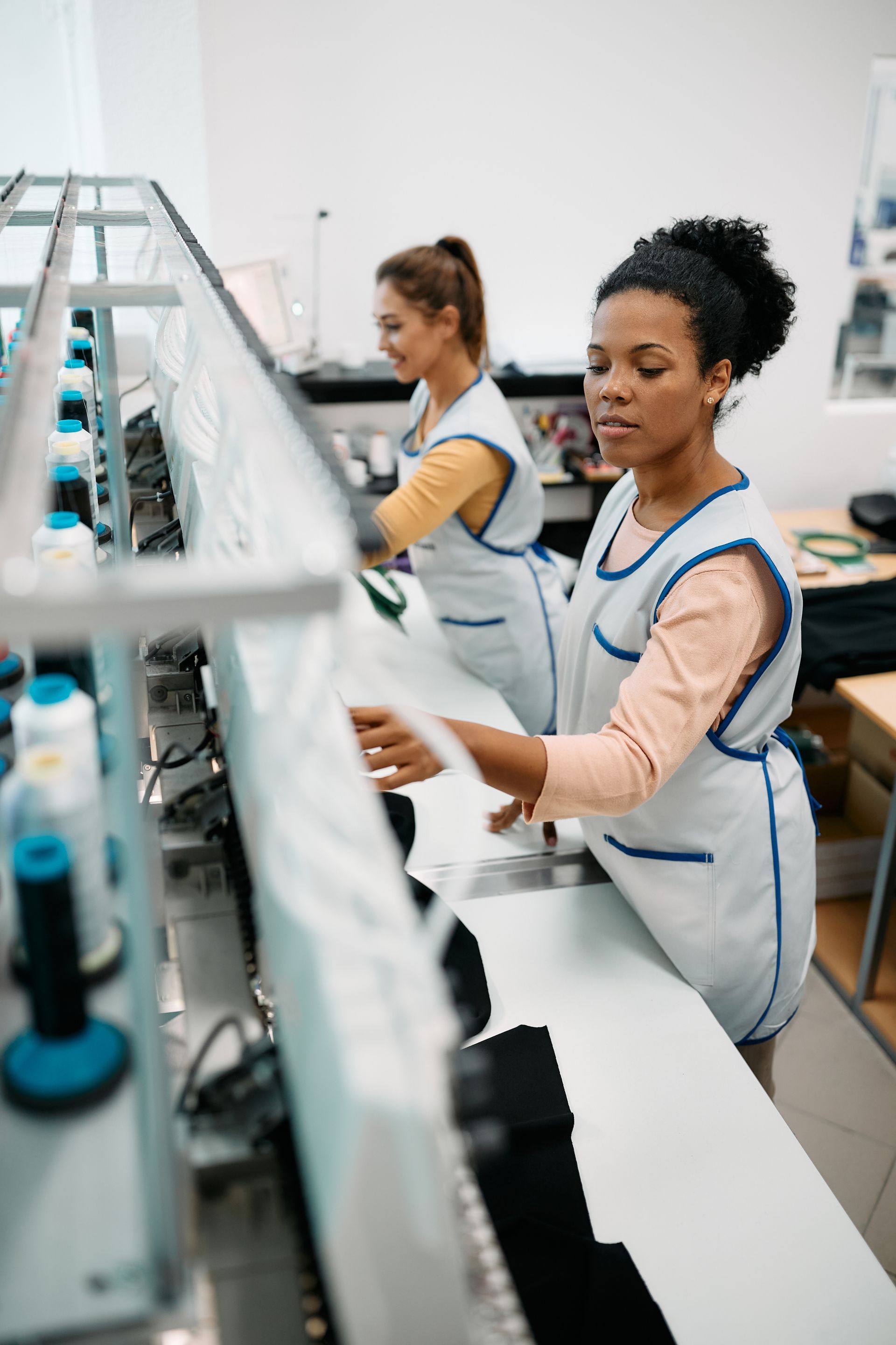 Two garment workers in light uniforms stand at an industrial table, focused on tasks at a sewing machine station.