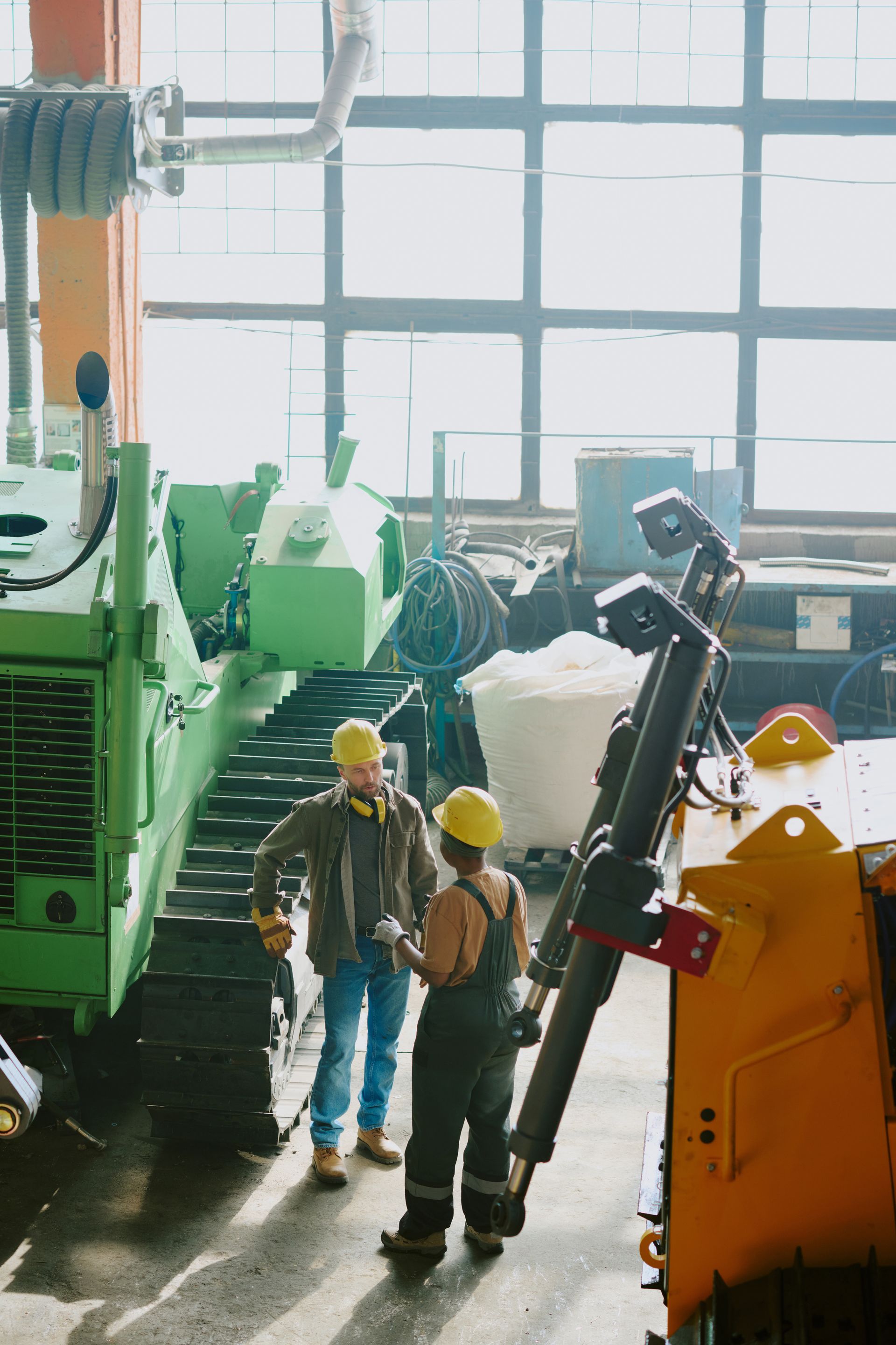 Two workers in hard hats stand between a green and a yellow piece of heavy machinery in an industrial warehouse.