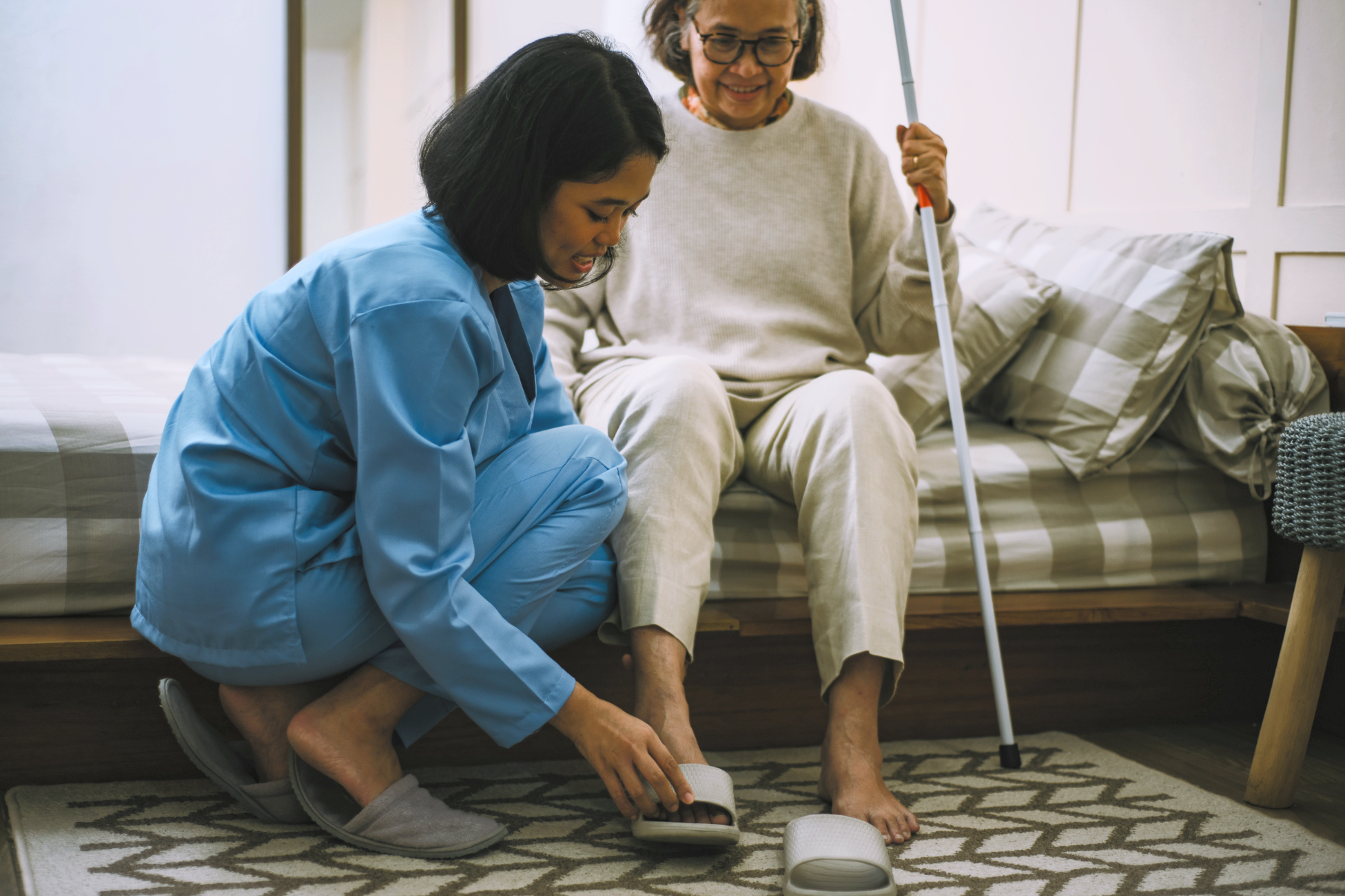 A caregiver assists an elderly woman, seated on a bed with a cane, by helping her put on a slipper.