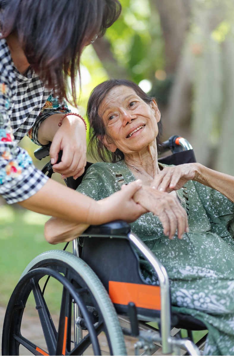 A young woman assists an elderly woman in a wheelchair outdoors. The elderly woman smiles.