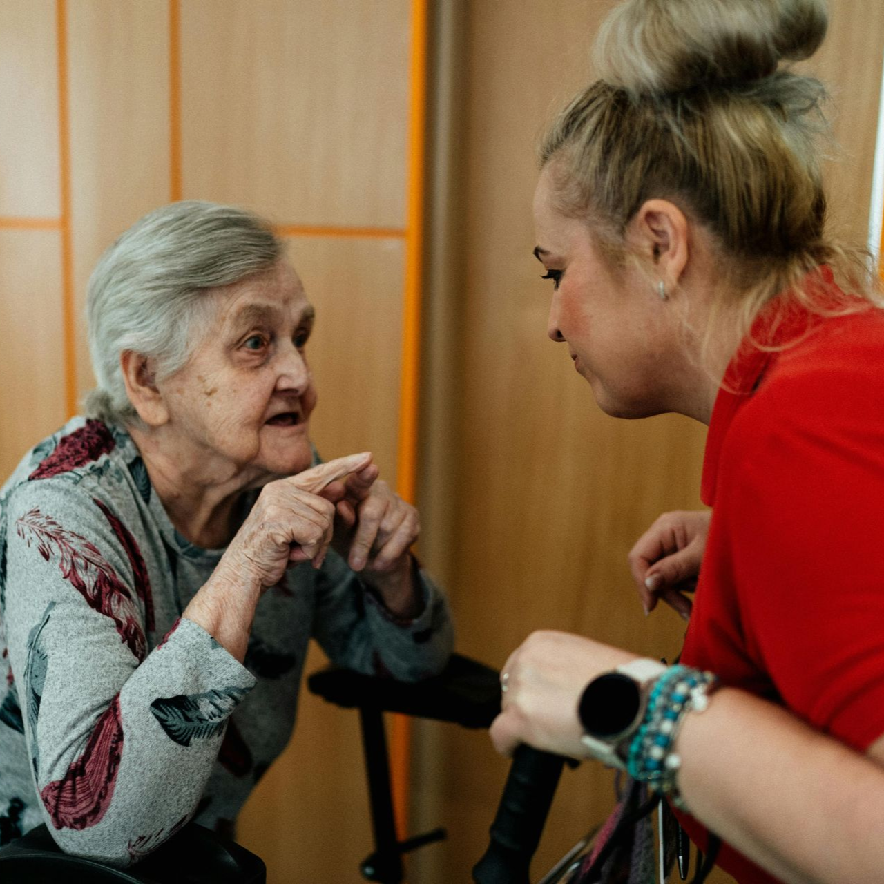 An elderly woman in gray shirt talking animatedly to a younger woman in red top indoors.