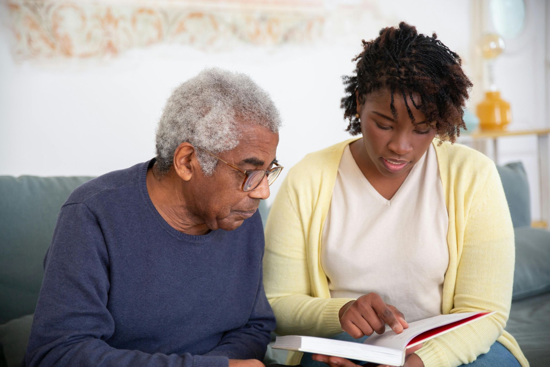 An older Black man and a younger Black woman read a book together on a couch indoors. The woman points to the pages.