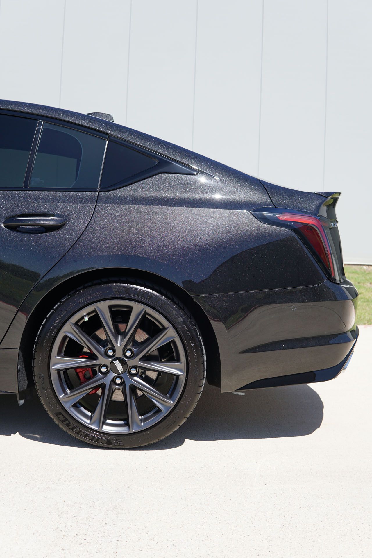 Dark gray Cadillac sedan's rear quarter panel. Gray wheels, tinted windows, black spoiler, red brake calipers.