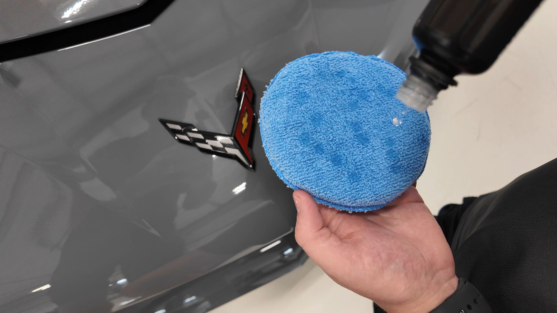 Person applying product to a blue buffing pad on a gray car next to the Corvette emblem.