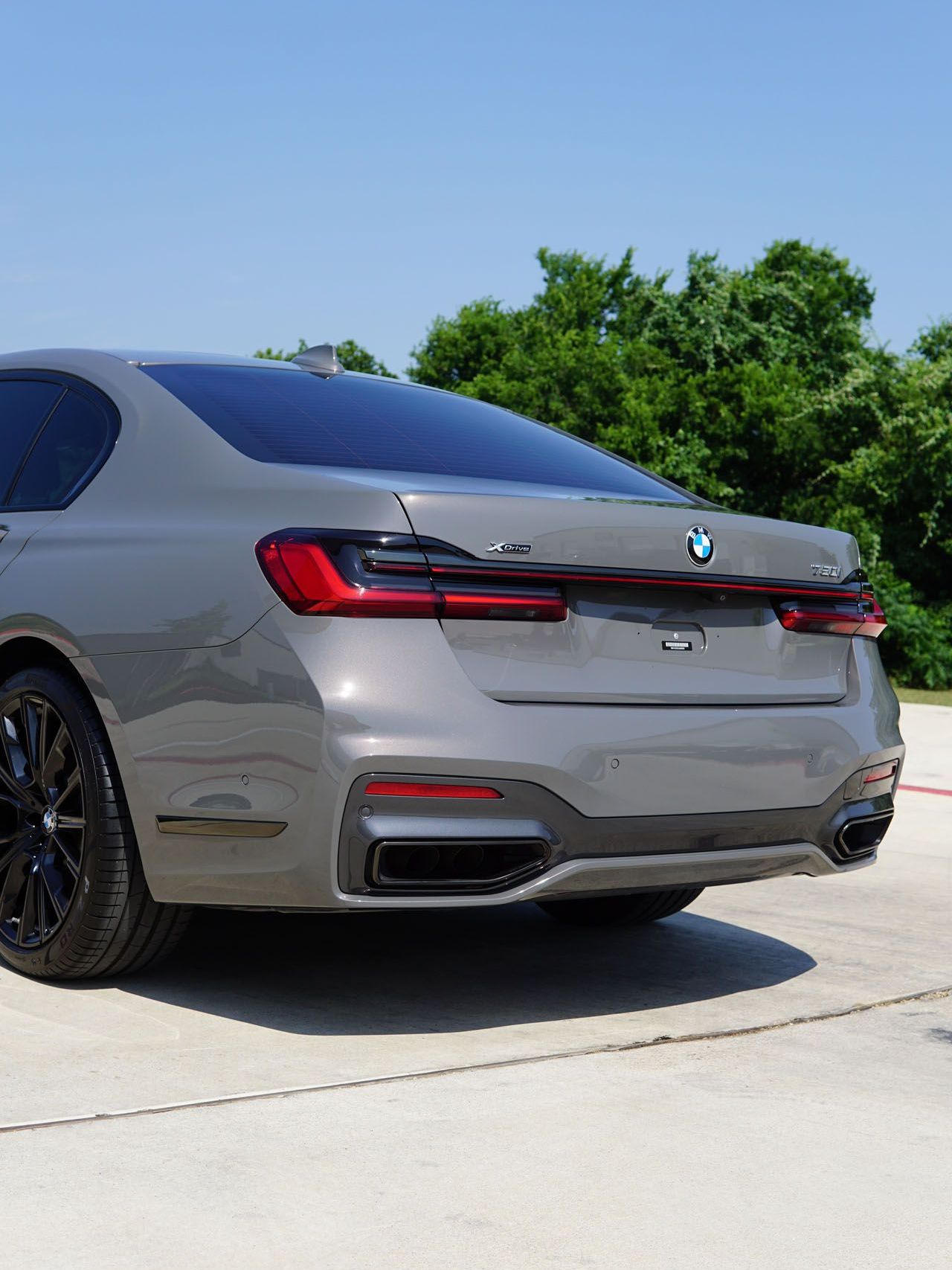 Gray BMW 7 Series sedan, rear view, black wheels, parked on concrete, trees in background.