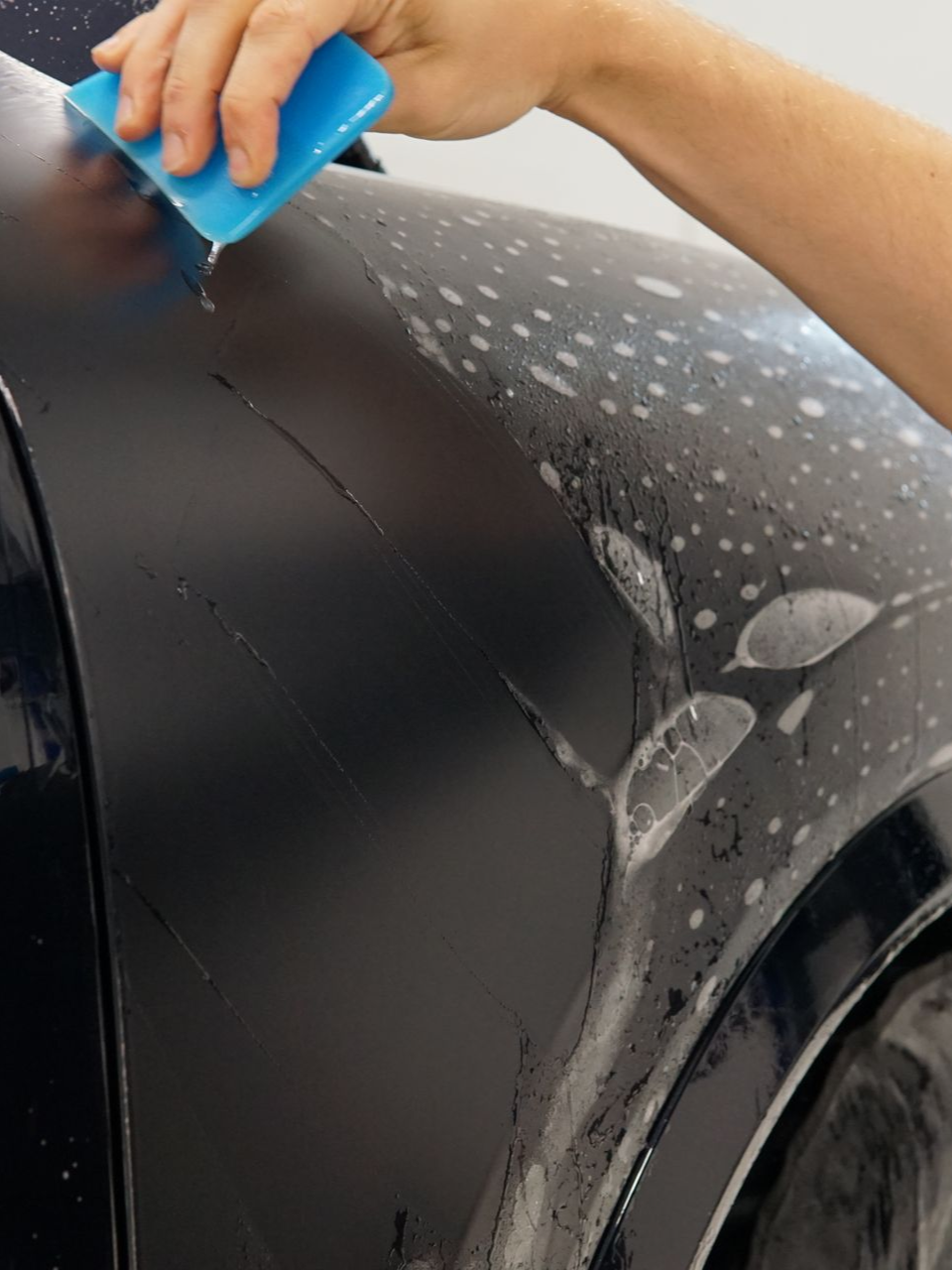 Person applying film to a black car fender, using a blue squeegee.
