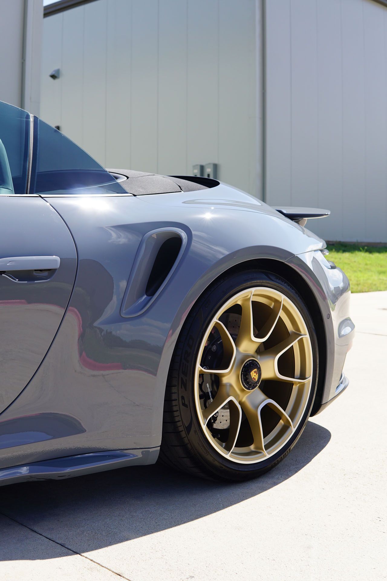 Gray Porsche convertible with gold wheels, parked on asphalt near a building.