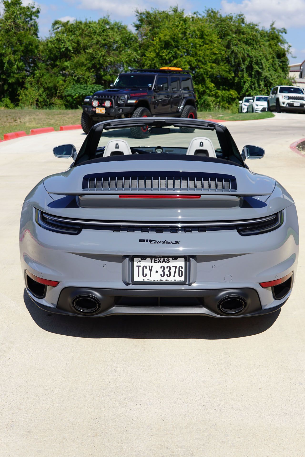 Gray Porsche convertible with top down, Texas license plate, parked on asphalt. Black Jeep SUV in background.