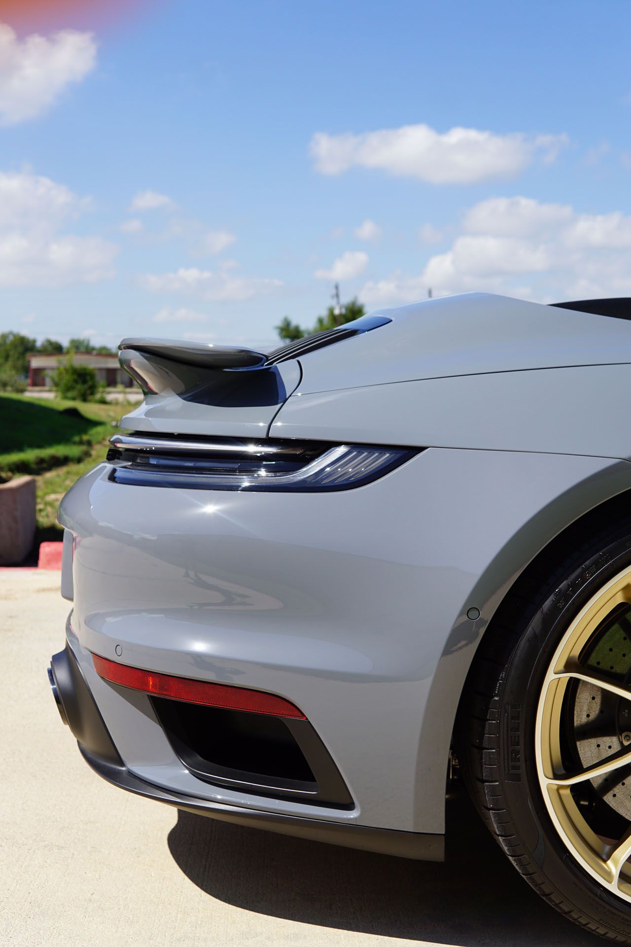 Rear quarter view of a light gray Porsche sports car with gold wheels, spoiler, and red taillight against a blue sky.