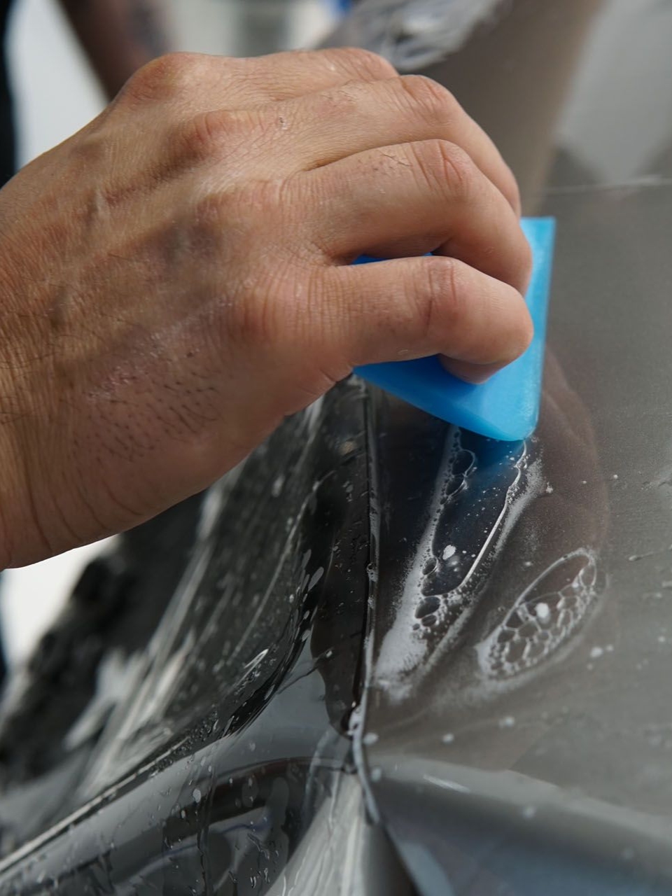 Hand smoothing out protective film on a car surface with a blue squeegee.