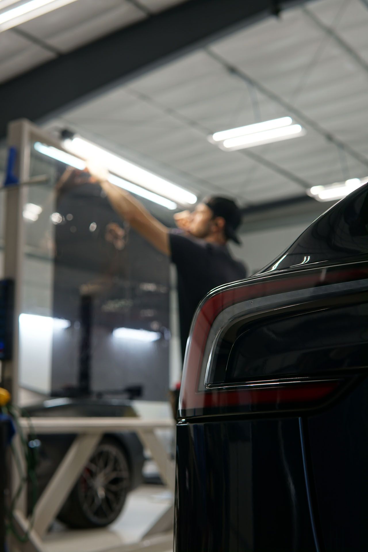Person applying tint to a car window inside a shop. Tail light in focus, person in background.
