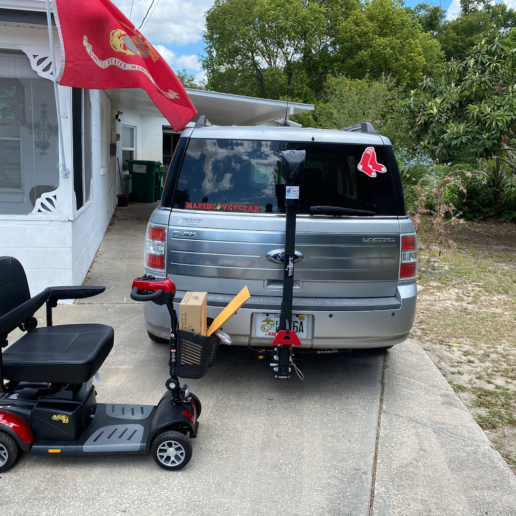 Silver SUV with a mobility scooter hitch carrier, parked in a driveway beside a house with a red flag.