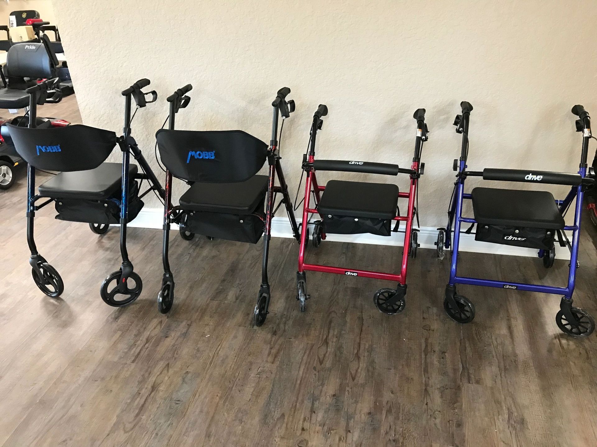 Four rollator walkers in black, red, and blue lined up on a wood floor indoors.