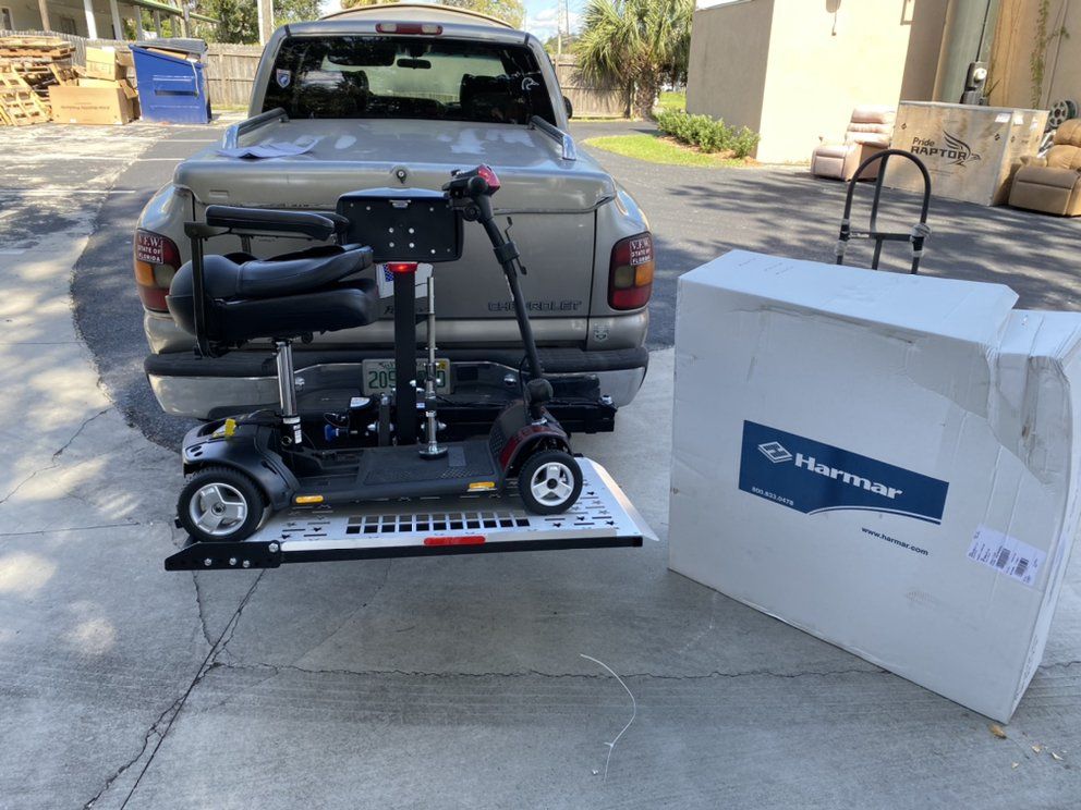Wheelchair scooter loaded on a trailer behind a pickup truck, with a large white box beside it.