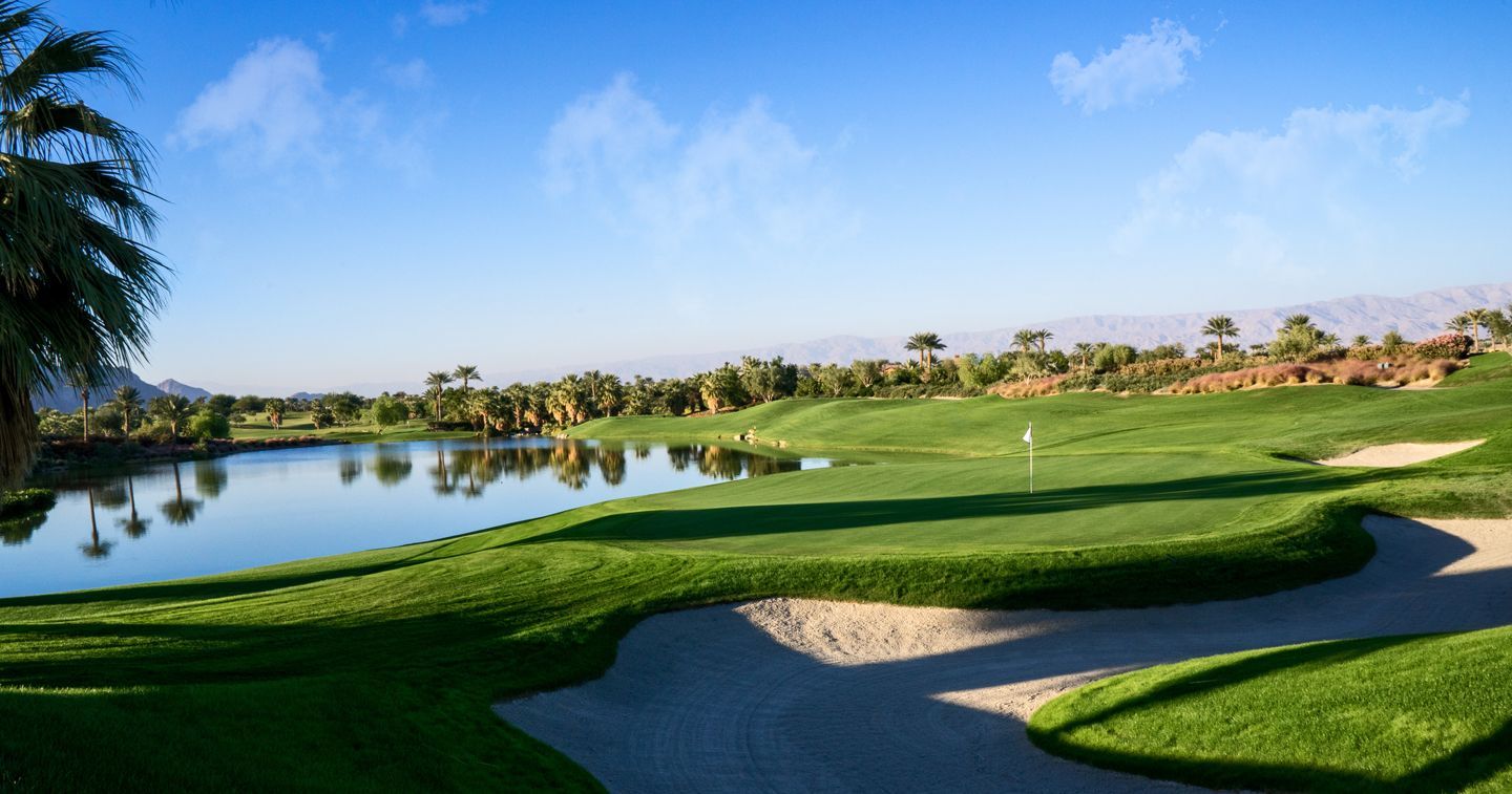 A sunny golf course with a large pond on the left, a green putting area with a flag, and sandy bunkers under a clear sky.