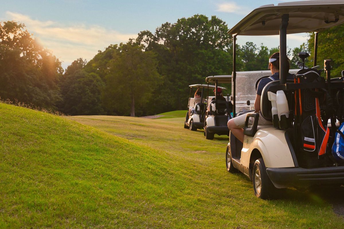 Three golf carts lined up on a grassy course at sunset, with a person seated in the foreground cart.