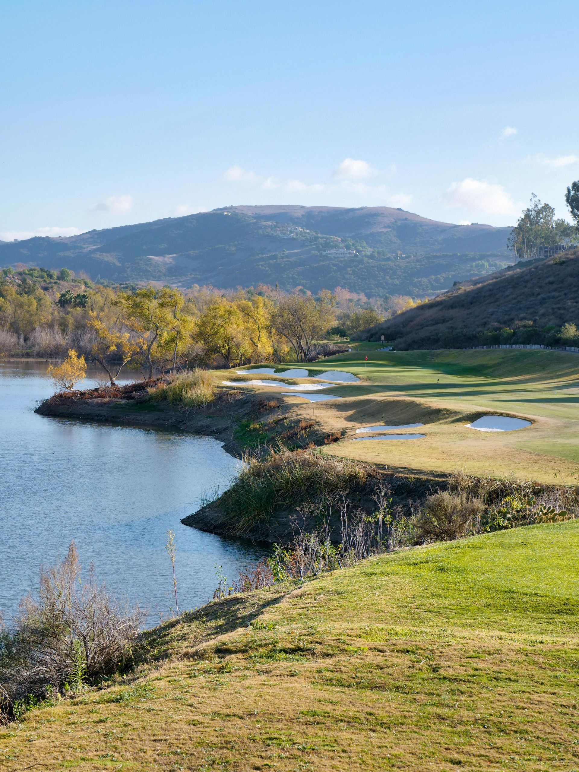 A sunlit golf course hole curves along a calm lake, bordered by grassy banks, sand traps, and rolling hills under blue sky.