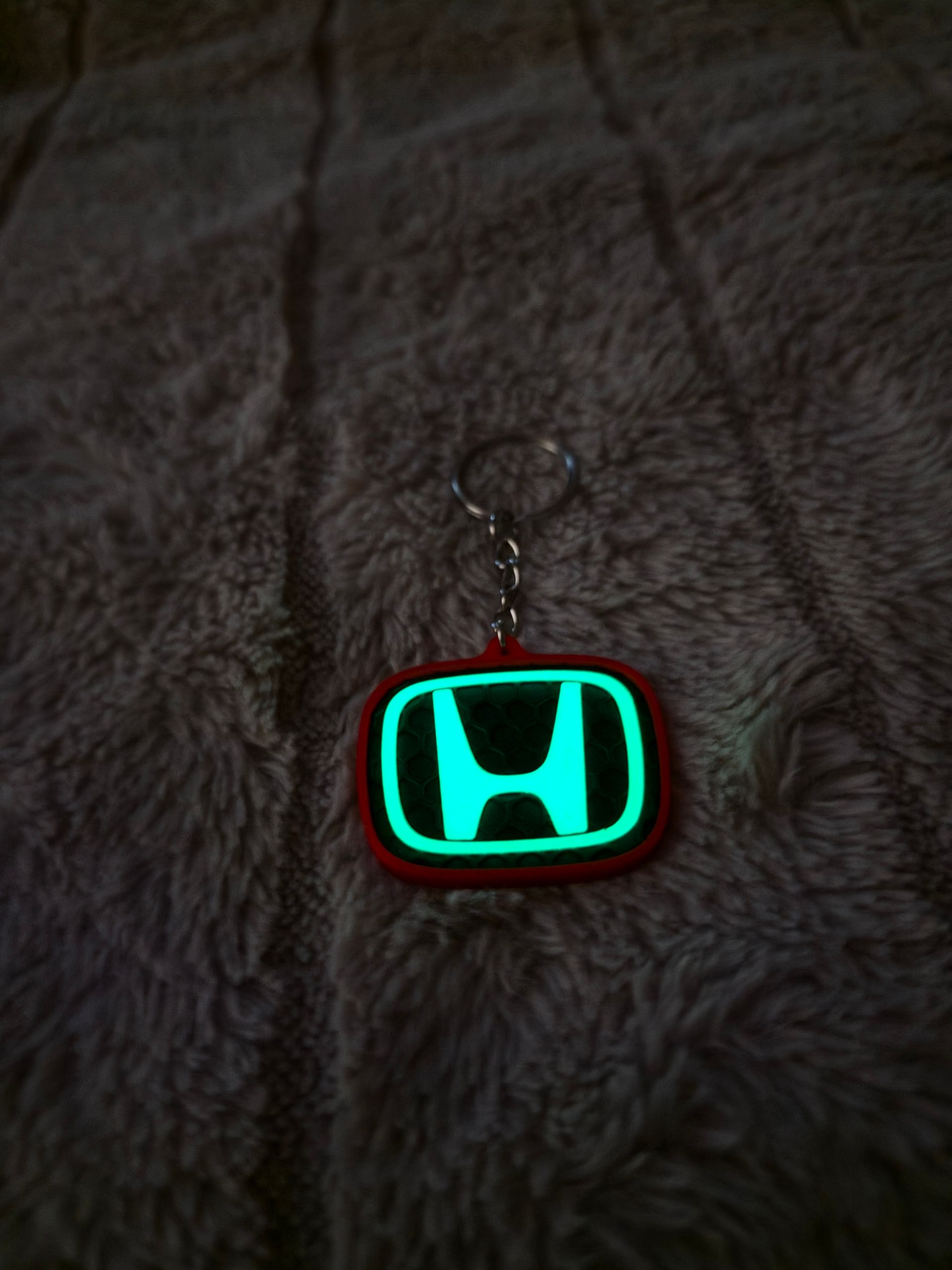 Sleutelhanger met Honda-logo en turquoise neon omlijning op een donkere stoffen achtergrond.