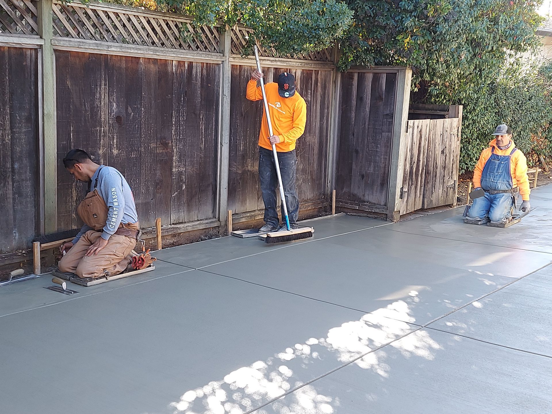 Three workers in work clothes smooth a newly poured concrete patio alongside a wooden fence.