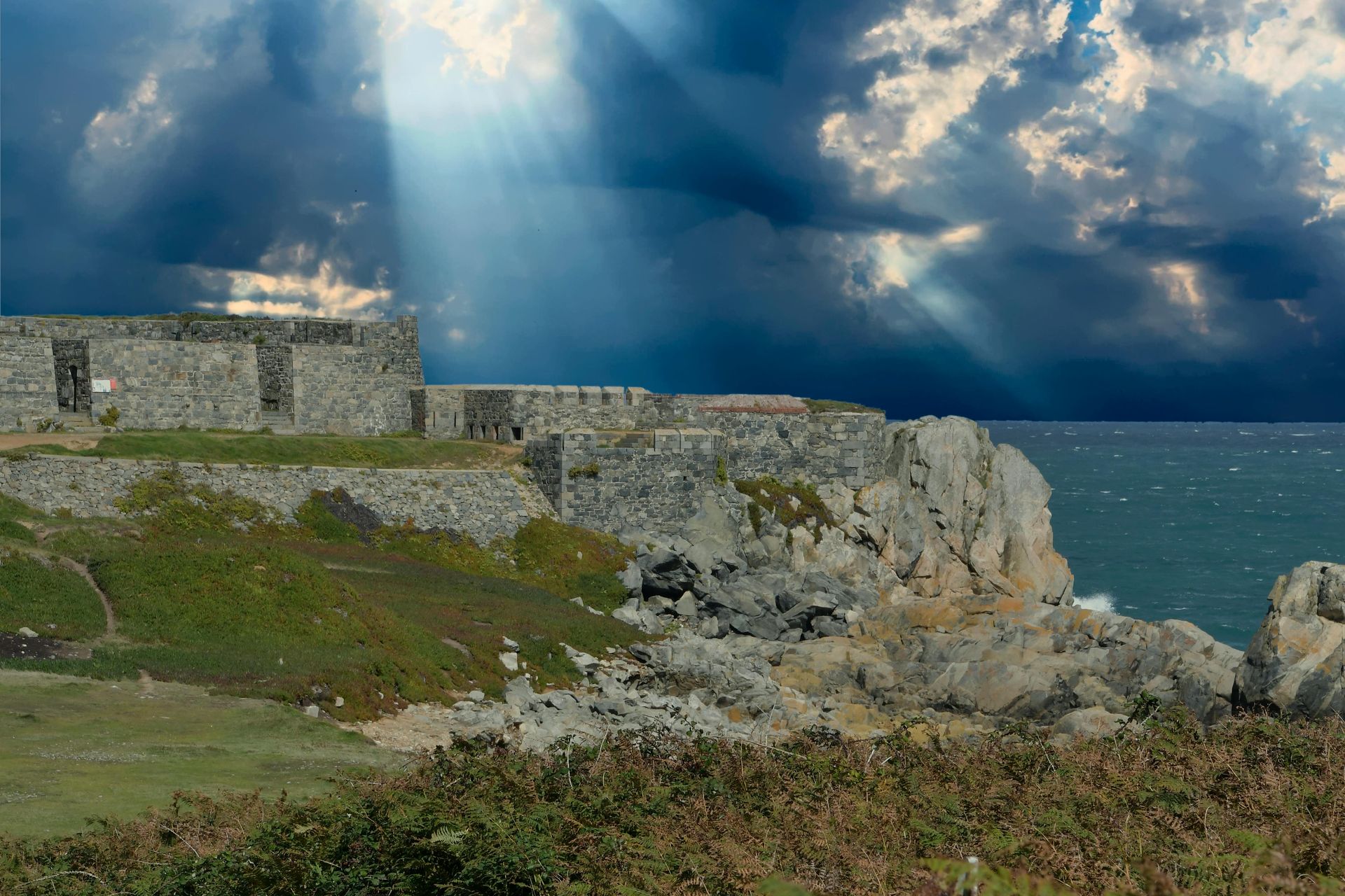 Stone ruins on a cliff overlooking the sea under a cloudy sky with sunbeams.