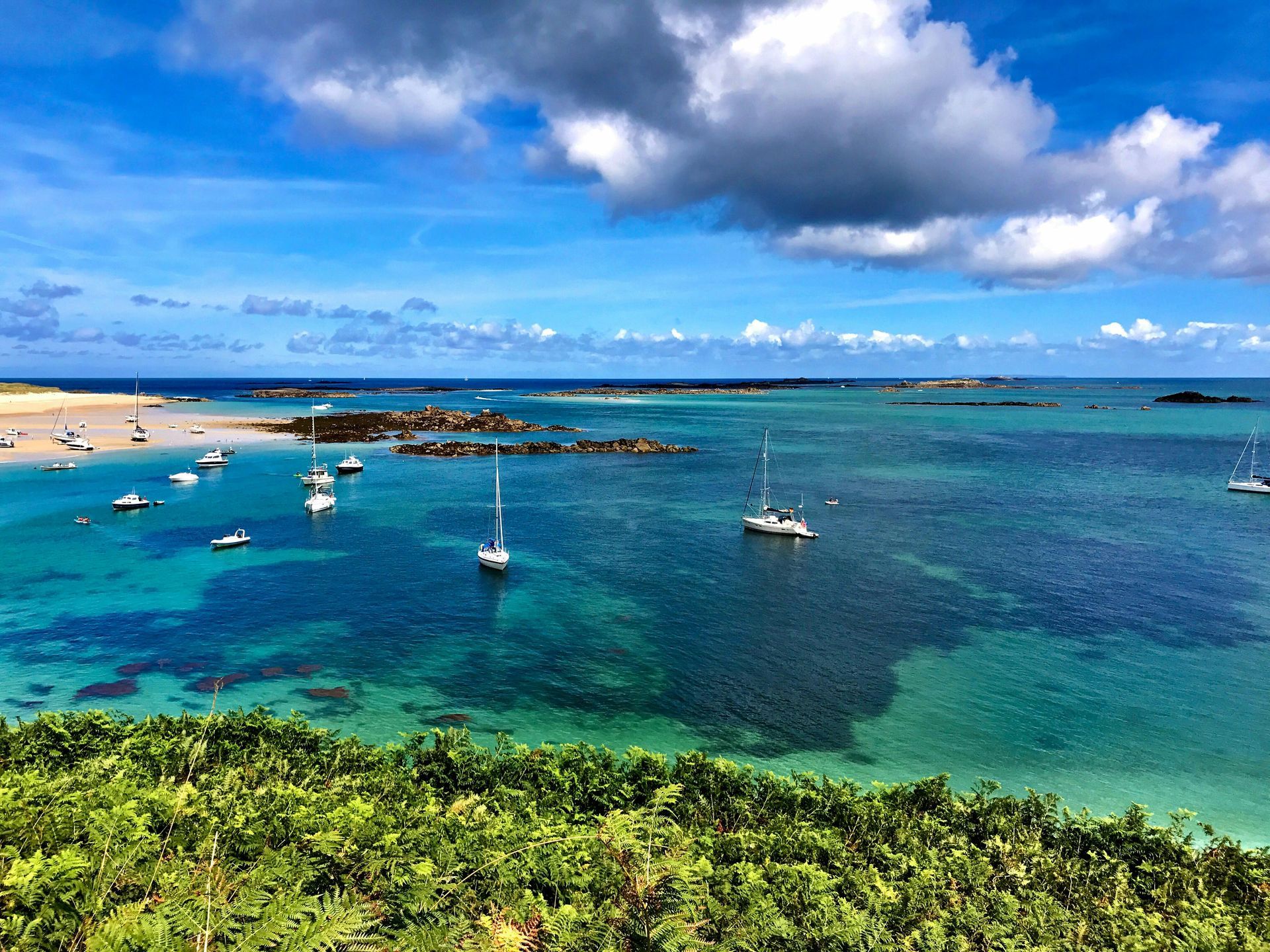Bright blue ocean with boats and a cloudy sky, seen from a green hillside.