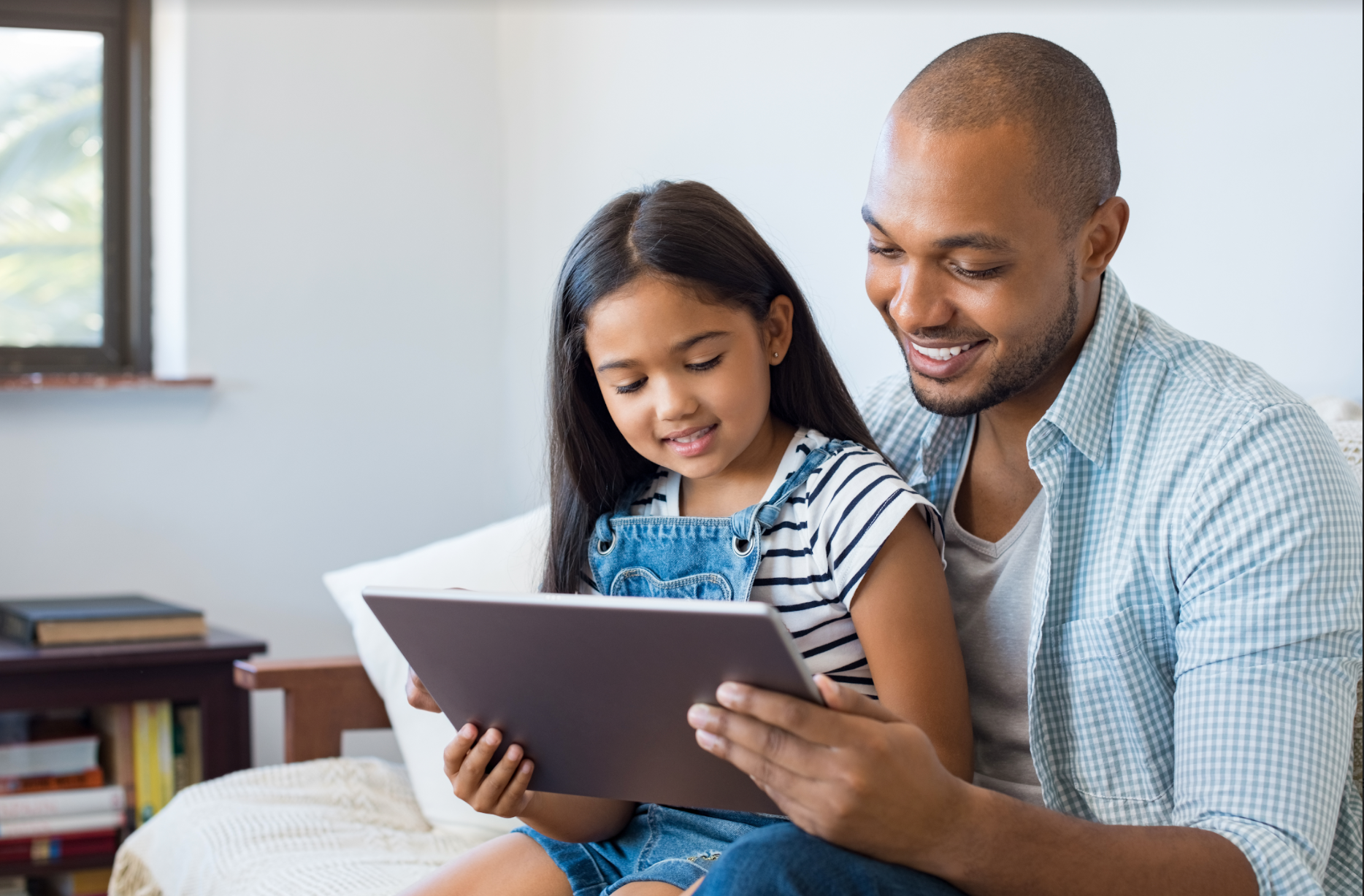 A man and a little girl are sitting on a couch looking at a tablet.