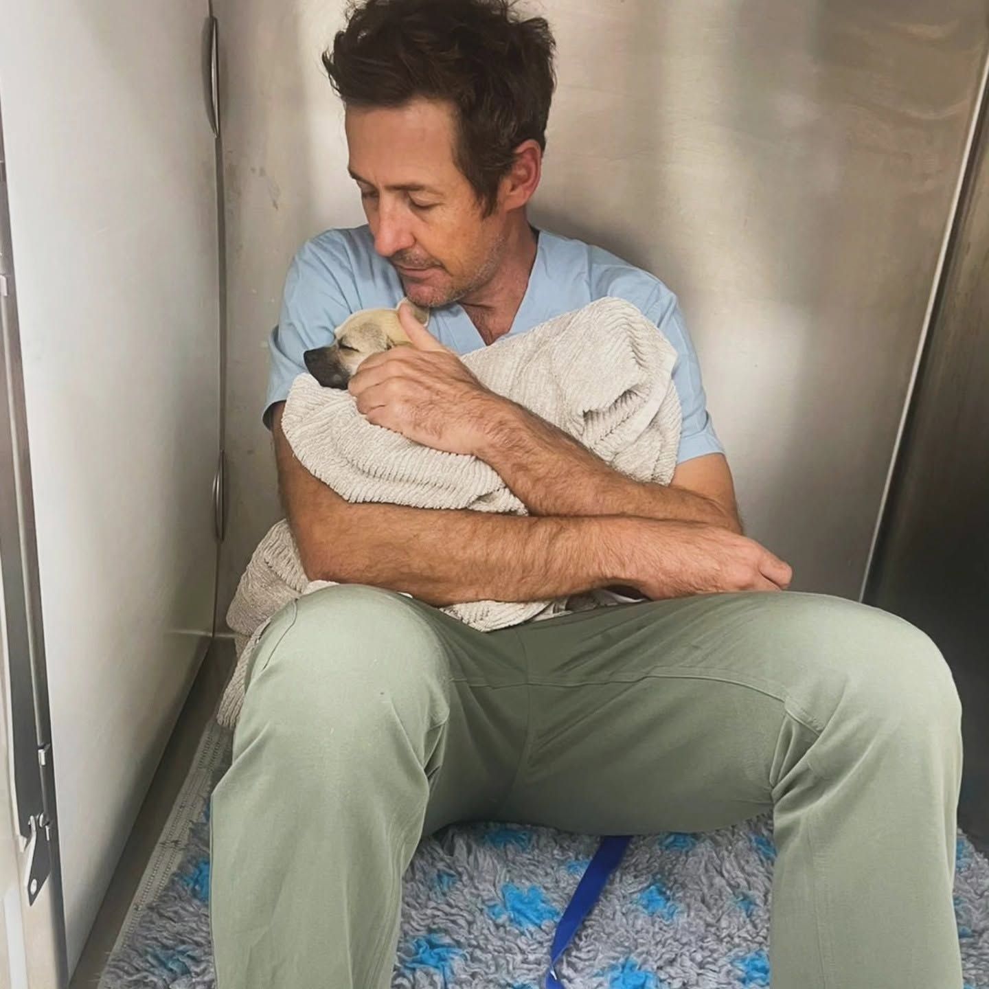 Man seated on the floor, cradling a bundled baby against a metal wall.