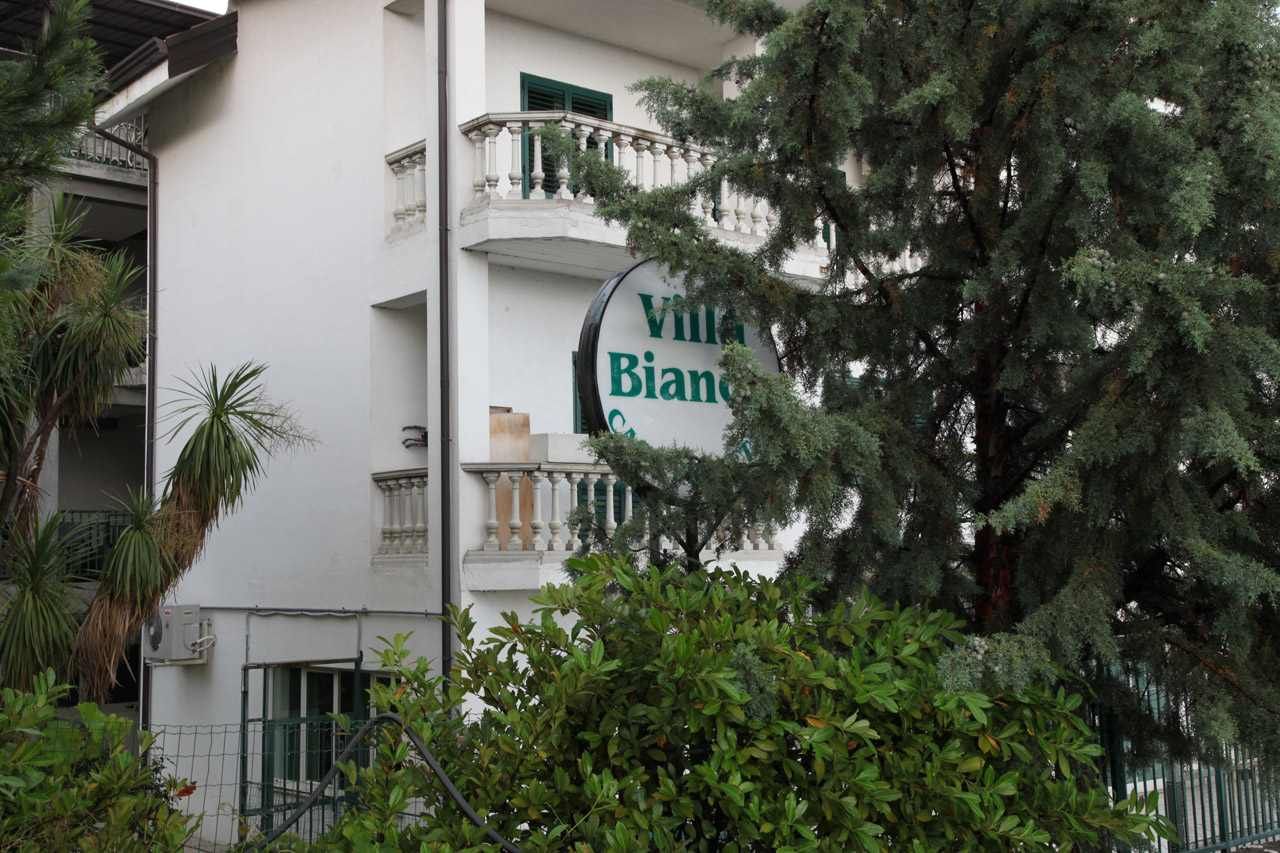 Edificio alberghiero bianco, parzialmente nascosto dagli alberi, con un'insegna sul balcone che recita 