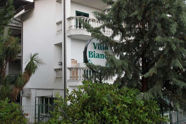 Edificio alberghiero bianco, parzialmente nascosto dagli alberi, con un'insegna sul balcone che recita