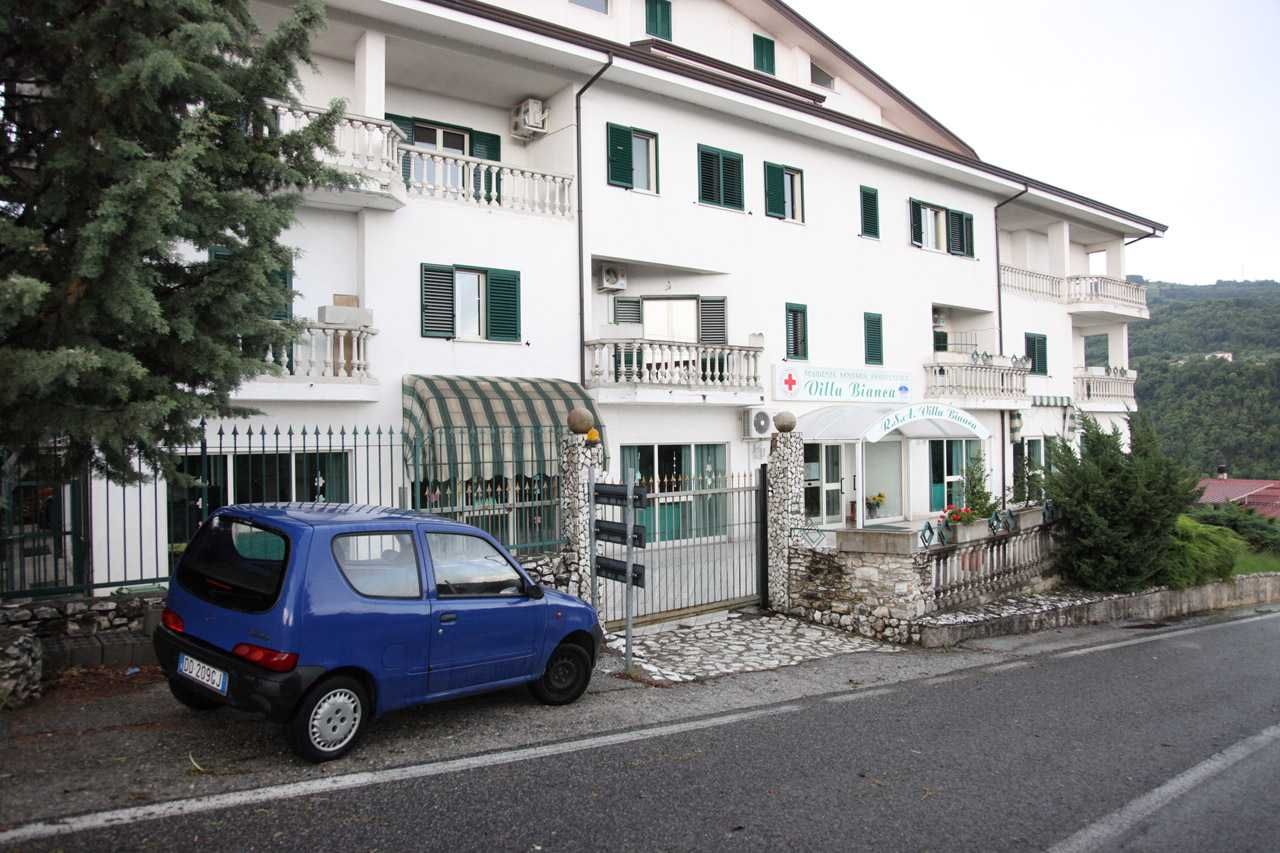 Un'auto blu parcheggiata davanti a un edificio bianco con persiane verdi in una strada tranquilla.