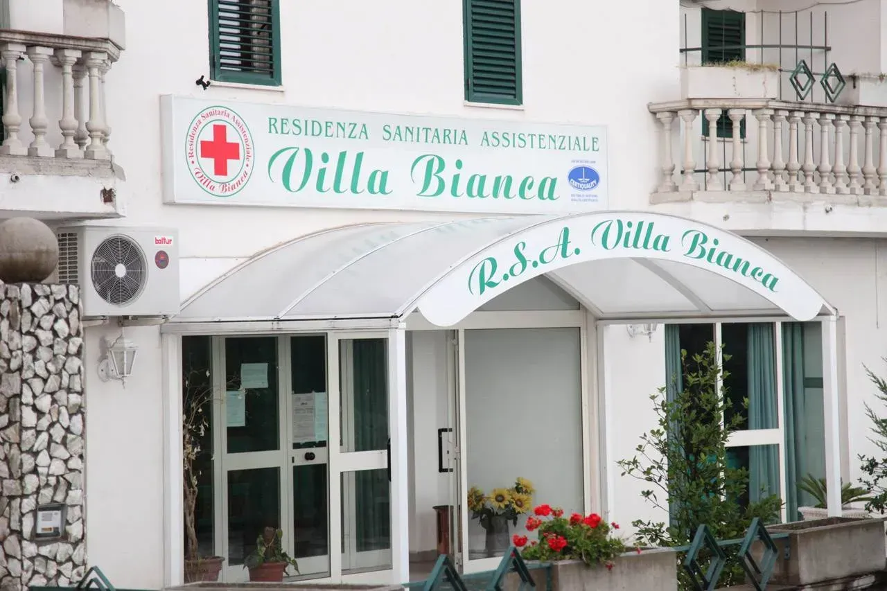 Facciata della clinica con tenda da sole verde, insegna a croce rossa e fioriere su un edificio bianco.
