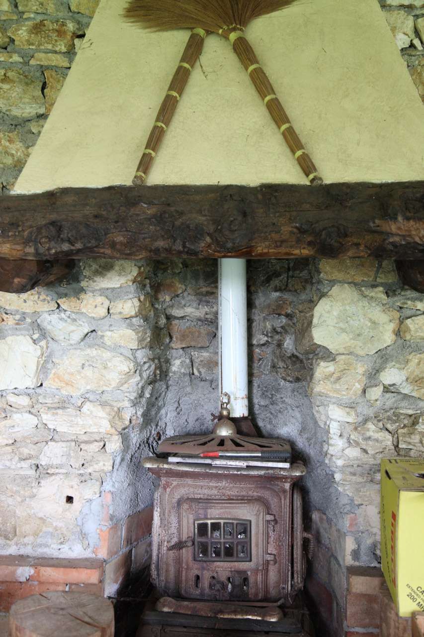 Camino rustico in pietra con stufa a legna e canna fumaria bianca sotto una trave di paglia.