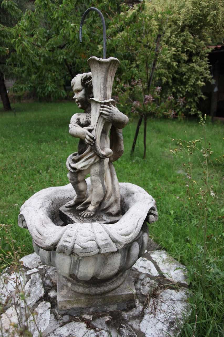 Fontana da giardino in pietra con figura scolpita e vasca, circondata da erba e alberi.