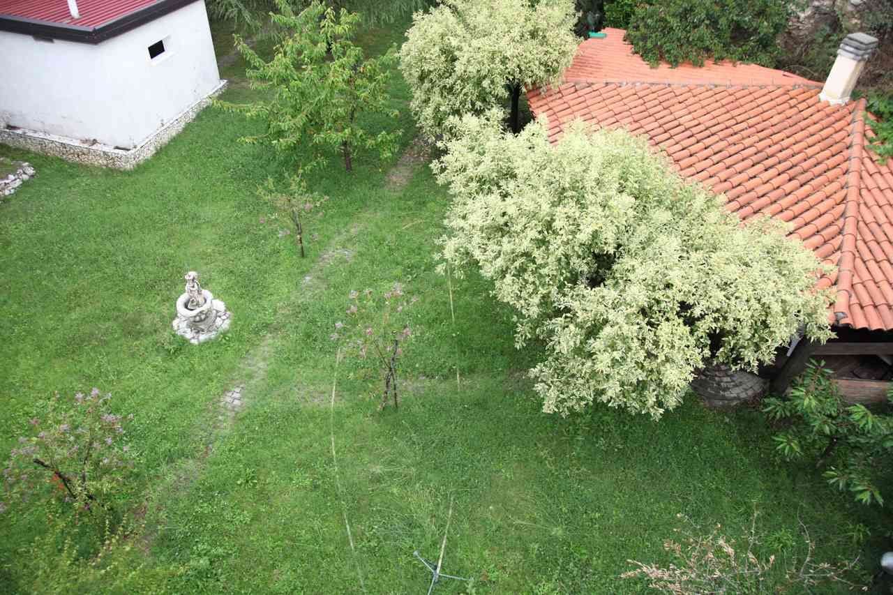 Veduta aerea di un prato con una fontana bianca, alberi frondosi ed edifici con tetti rossi.