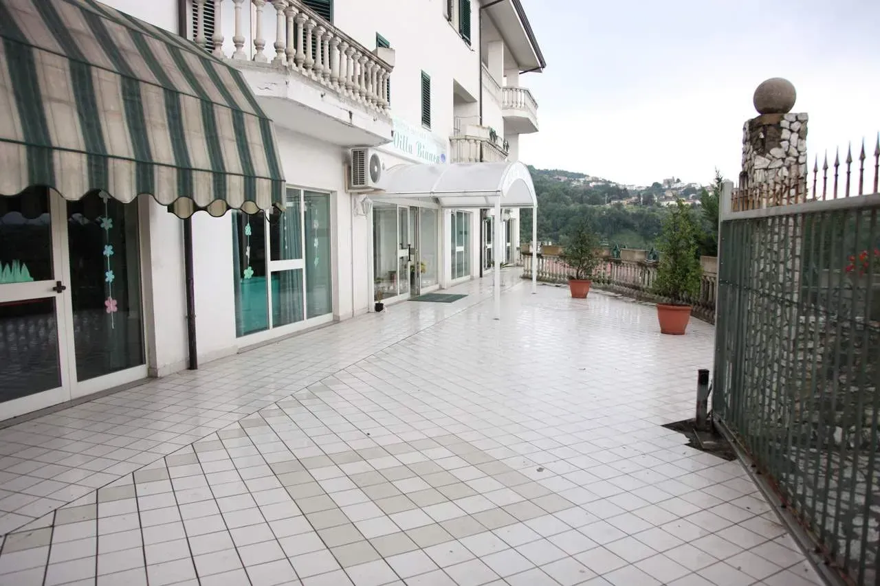 Terrazza piastrellata vuota accanto a edifici bianchi, con tenda da sole a strisce verdi e vista mare oltre il cancello.