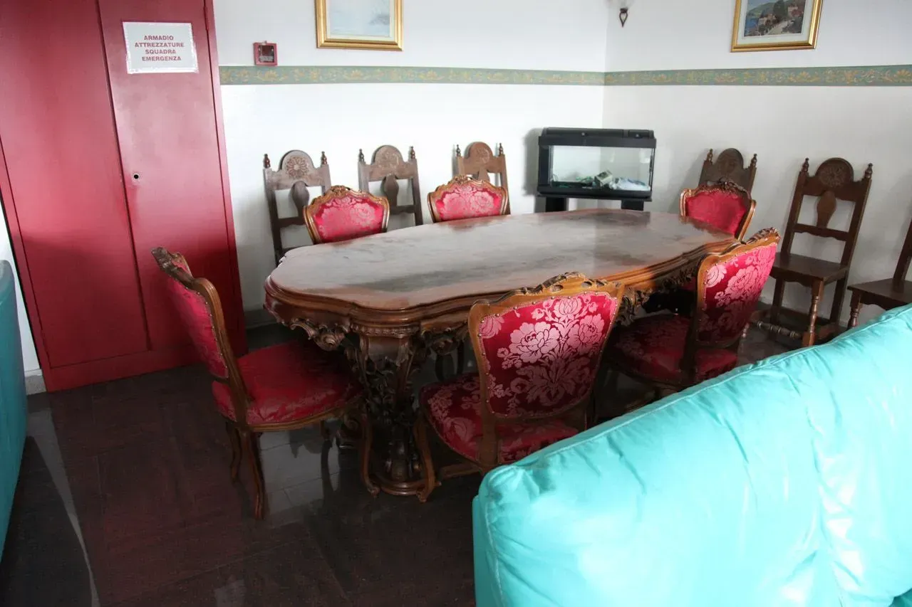 Sala da pranzo con un lungo tavolo, sedie rivestite di tessuto rosso e un televisore appoggiato alla parete di fondo.