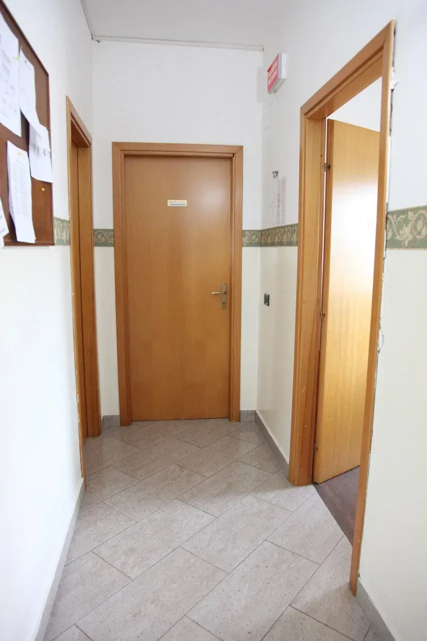 Stretto corridoio con pareti bianche, pavimento in piastrelle beige e porte di legno in fondo a destra.