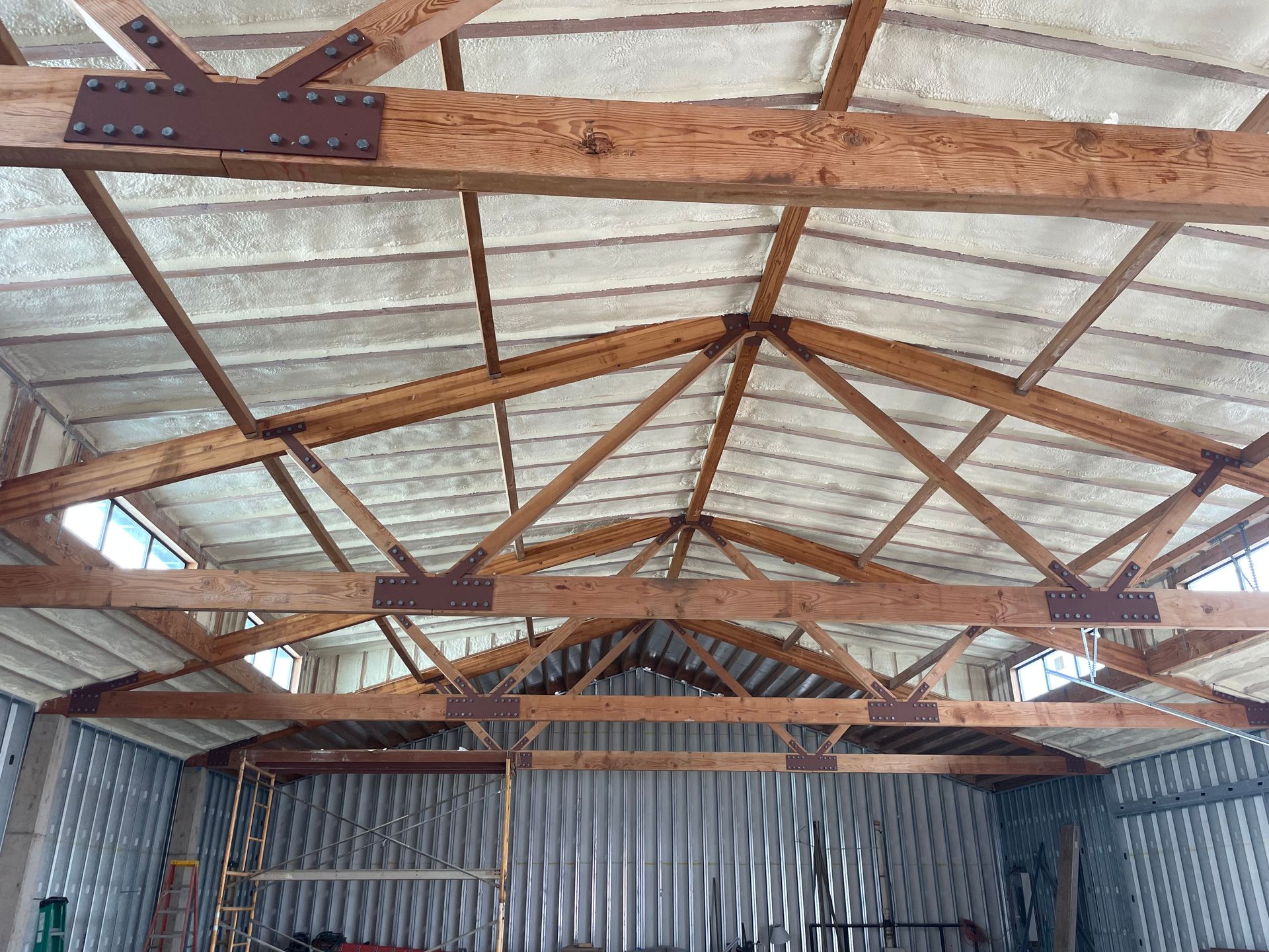 Interior view of a building's roof with wooden trusses and spray foam insulation against corrugated metal walls.